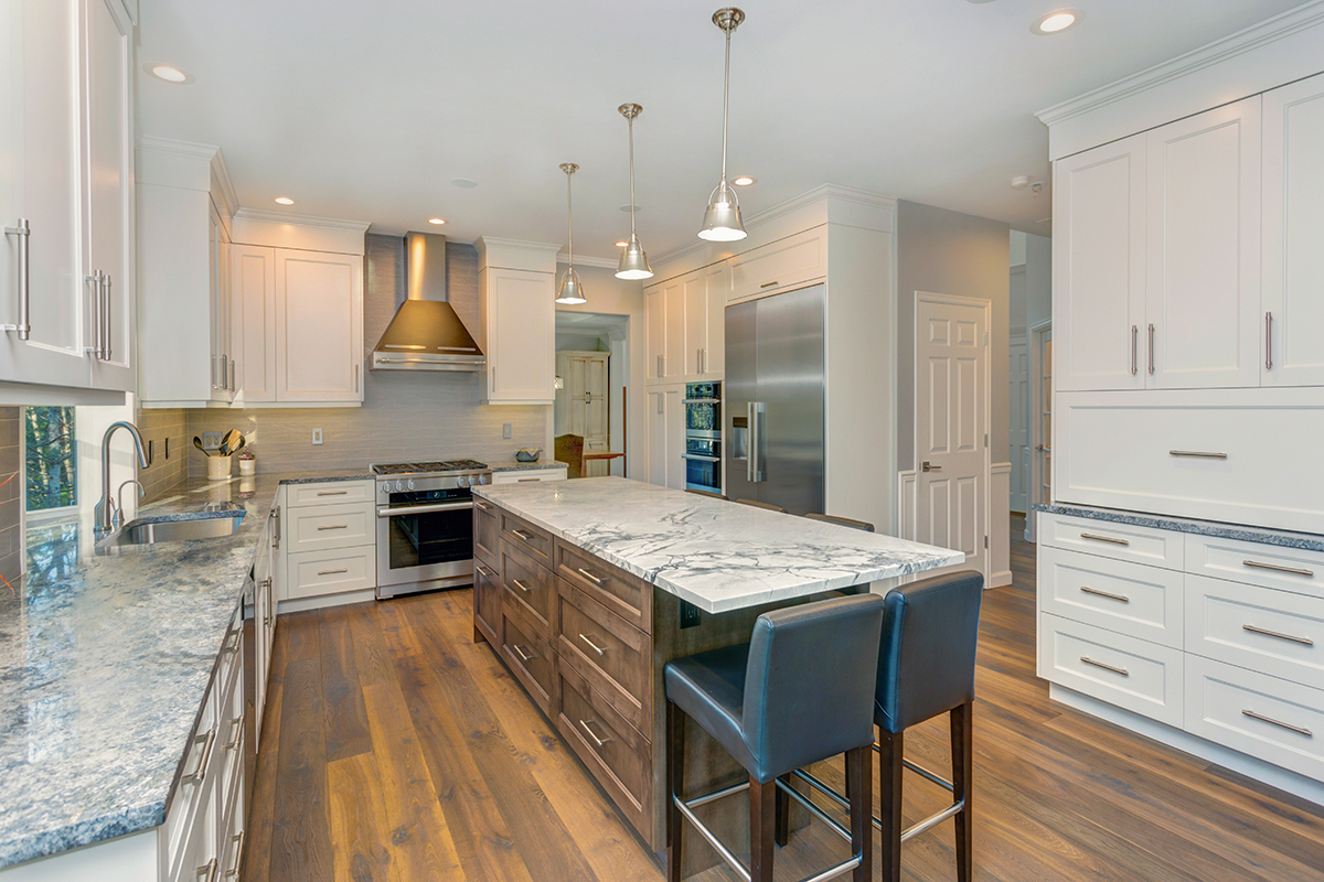 Luxury home interior boasts Beautiful black and white kitchen with custom white shaker cabinets, endless marble topped kitchen island with black leather stools over wide planked hardwood floor.